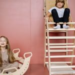 Two kids enjoying their time playing on a seesaw and a wooden climbing ladder indoors.